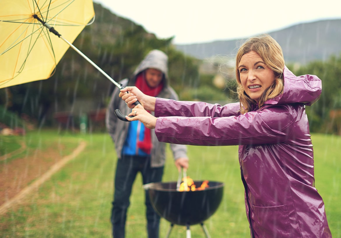 Natural Pharmaceuticals Imuberin® Esőben sárga esernyőt tartó nő lila esőkabátban, háttérben férfi esőben szabadtéri grillezés közben.